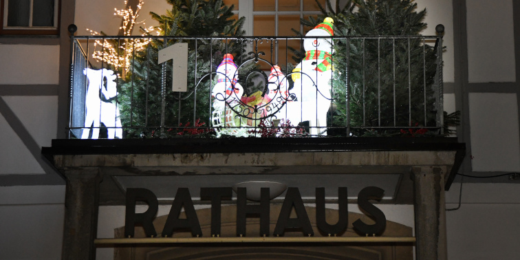 Weihnachtlich geschmückter Rathausbalkon mit Tannen, leuchtenden Figuren und der Zahl 1 – Teil eines lebendigen Adventskalenders