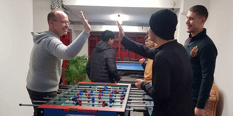 Mehrere junge Männer spielen Tischfußball und geben sich lachend High-Fives. Im Hintergrund stehen weitere Personen an einem Tischtennistisch. Die Stimmung ist locker und fröhlich.