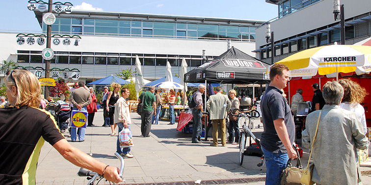 Menschen schlendern über einen sonnigen Außenmarkt mit mehreren Verkaufsständen und Firmenzelten.