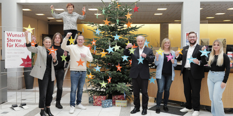 Acht Personen stehen vor einem Weihnachtsbaum in einer Bankfiliale, geschmückt mit bunten Sternen