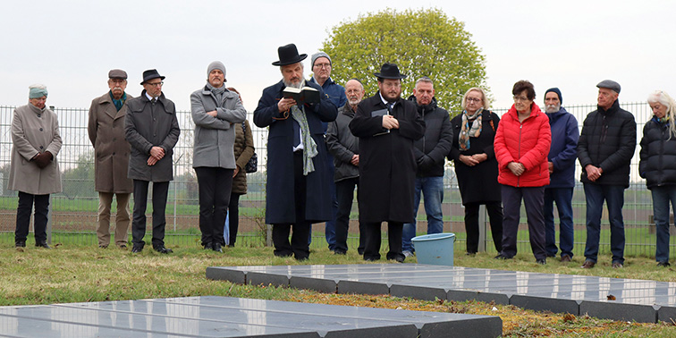 Eine Gruppe von Menschen steht auf einem Friedhof zusammen und nimmt vor mehreren Grabsteinen an einer Gedenkzeremonie teil.