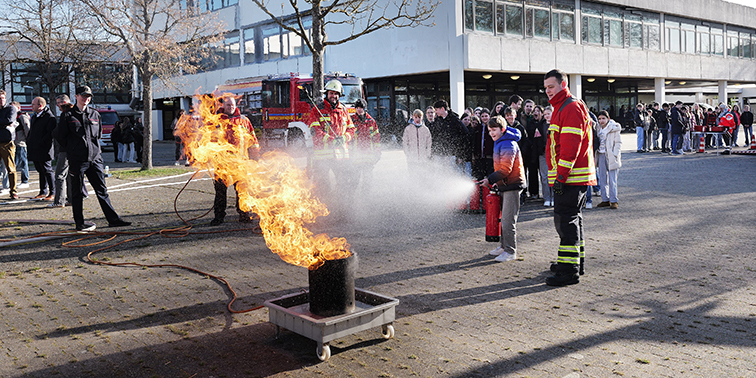 Kind löscht unter Anleitung der Feuerwehr ein offenes Feuer bei einer Vorführung im Freien. Feuerwehr bei einer Übung