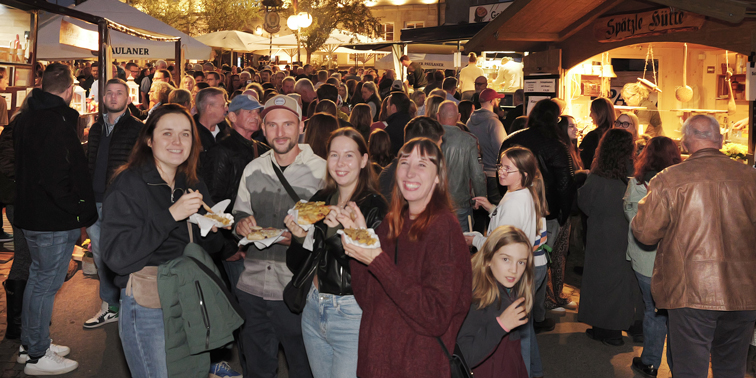 Gruppe lachender Menschen auf einem abendlichen Straßenfest, die Krautgerichte aus Papierschalen essen; im Hintergrund drängen sich Besucher vor beleuchteten Marktständen.