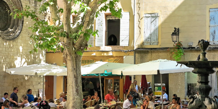 Gäste sitzen bei Sonnenschein in einem Manosquer Straßencafé (rechts: ein Brunnen)