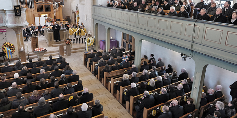 Große Trauerfeier in einer Kirche mit vielen sitzenden Gästen, Blumenkränzen und Urne vor dem Altar.