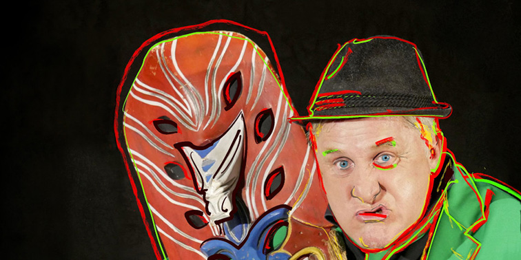 Ein Mann in grünem Sakko und Hut hält eine große, rot bemalte Holzmaske mit weißen und blauen Mustern vor dunklem Hintergrund.